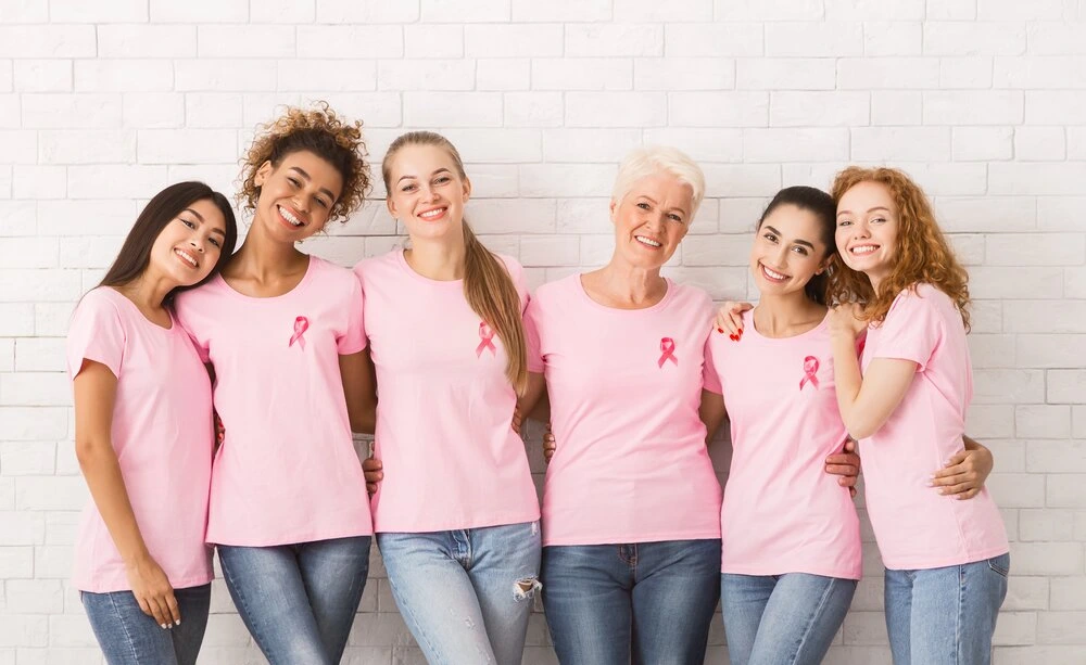 Happy Multiracial Ladies In Breast Cancer Pink T-Shirts With Awareness Ribbons Embracing Standing Near White Brick Wall Indoor