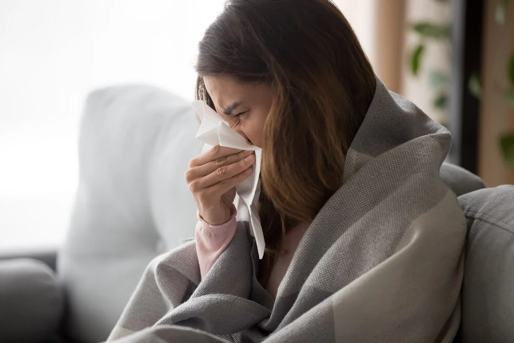 Ill upset young woman sitting on sofa covered with blanket freezing blowing running nose